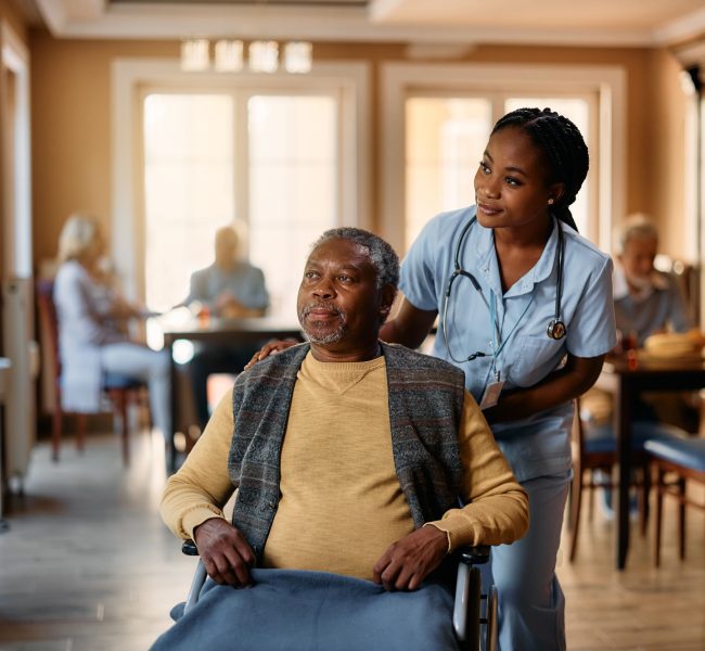 Black nurse and senior man in wheelchair looking through the window at nursing home.